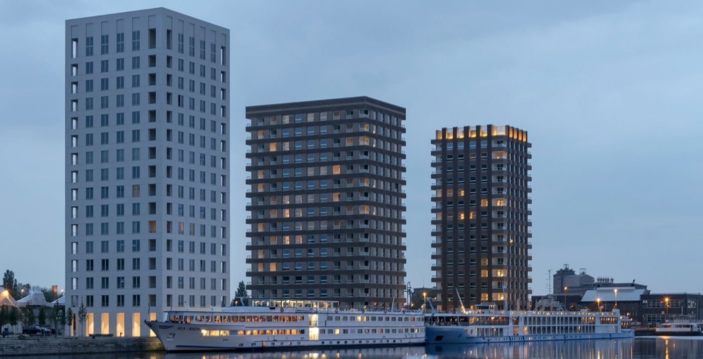 Drei moderne Hochhäuser erheben sich am Flussufer, beleuchtet in der Dämmerung