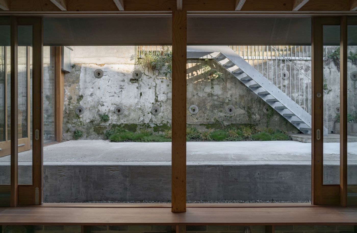 Ein Innenraum mit Holzbalken und Glaswänden blickt auf einen Betonhof mit Treppe.