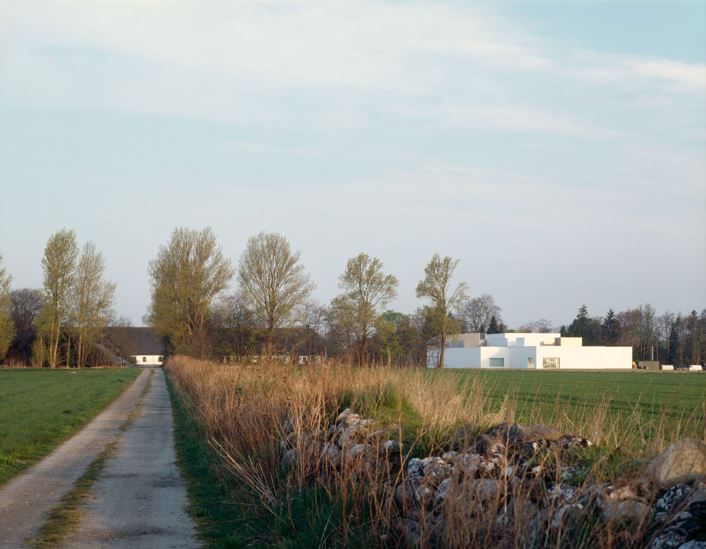 Ein weisses, kubisches Gebäude steht in einer grünen Landschaft neben einem Feldweg