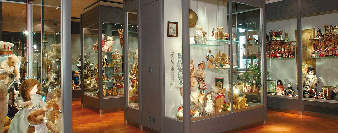 A room with glass cases displaying antique teddy bears, dolls, and miniature houses on wooden flooring