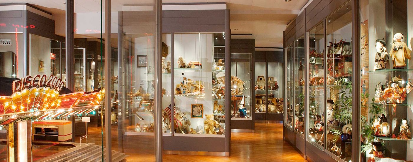 A colorful collection of teddy bears and dolls behind glass display cases in a well-lit room