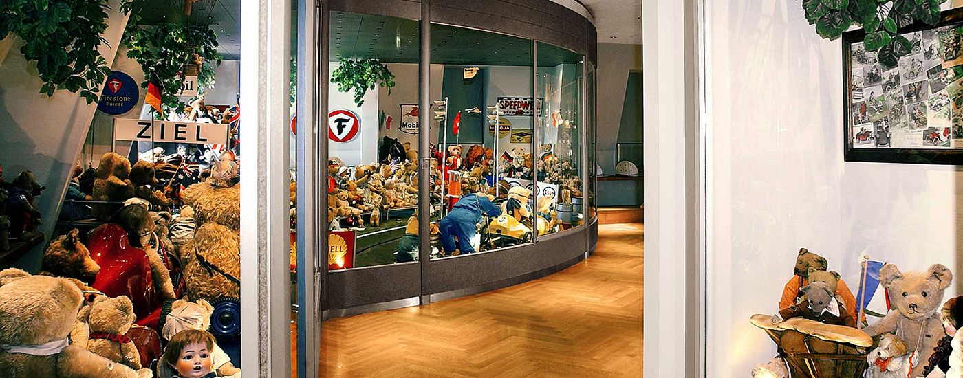 Teddy bears and dolls fill a colorful display case in an exhibition room with wooden flooring