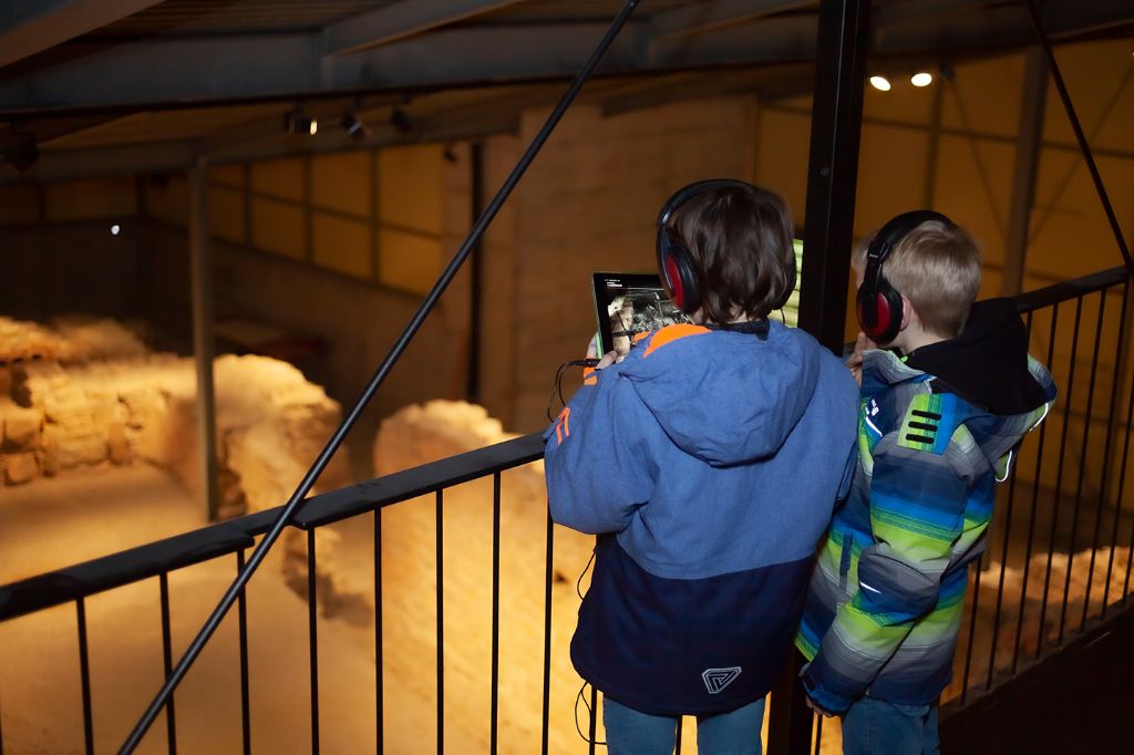 Zwei Kinder mit Kopfhörern erkunden eine historische Stätte mit einem Tablet