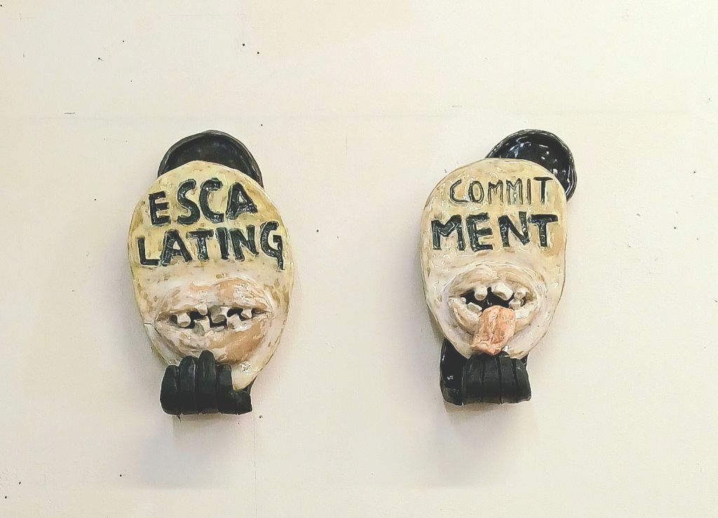 Zwei skulpturale Masken mit den Worten "Escalating" und "Commitment" hängen an der Wand