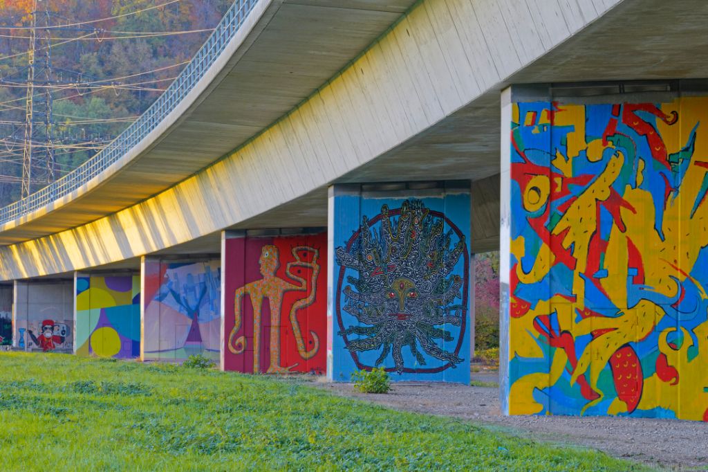 Under a bridge, colorful murals with abstract and figurative designs are displayed.