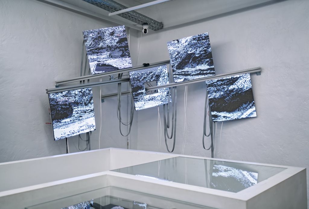 Sechs Bildschirme zeigen monochrome Pixelgrafiken in industrieller Galerieumgebung