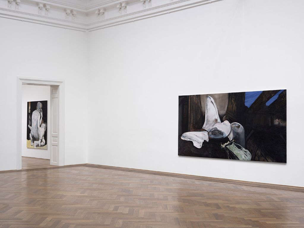 Two paintings in a white gallery room; one depicts abstract shapes, the other a figure.