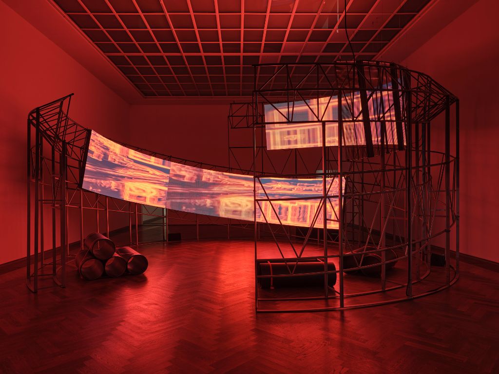 An abstract metal structure glows in red light, with video projections on screens