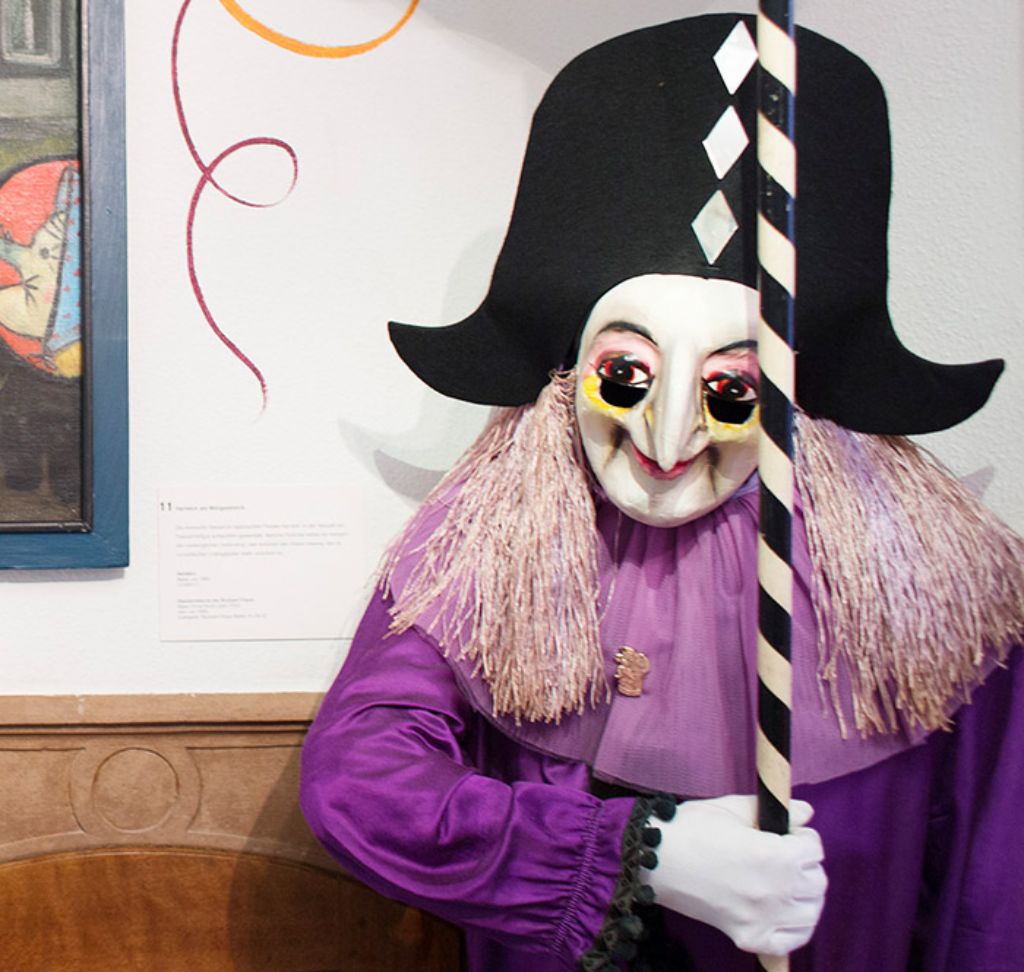 Auf einer Bank sitzt ein violett bekleideter Harlekin eine Fasnachtsfigur Er hält eine Stablaterne in den Händen