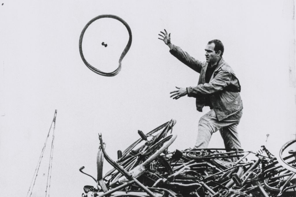 Ein Mann wirft ein grosses Rad in die Luft, während er auf einem Haufen alter Fahrräder steht.