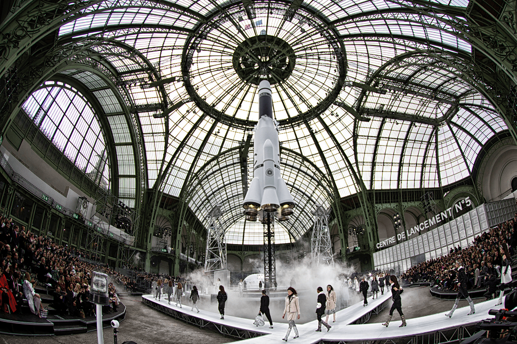 Models walk past a large rocket on a runway under a glass dome